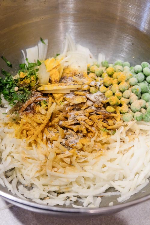 A bowl containing the grated potatoes and the spices, chopped onion, cilantro, and frozen peas, before mixing.