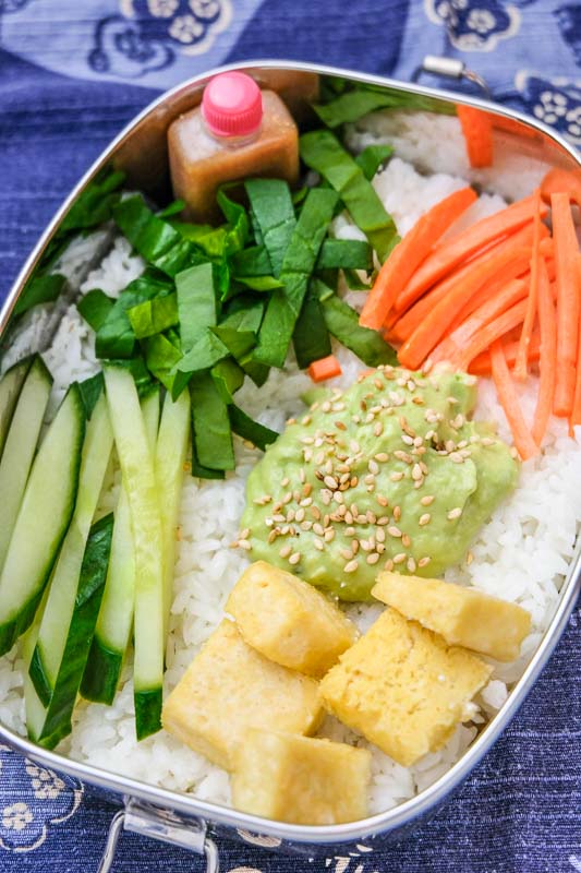 Vegan buddha bowl with baked tofu, cucumber, avocado, carrots, spinach, sesame seeds, and sesame dressing on rice, inside a stainless steel lunch box over a Japanese cloth wrapper. #veganrecipes #veganbuddhabowl #buddhabowlrecipes #vegandinners #veganlunch #healthyveganrecipes #buddhabowl #vegandiet #tofurecipes #veganmeals