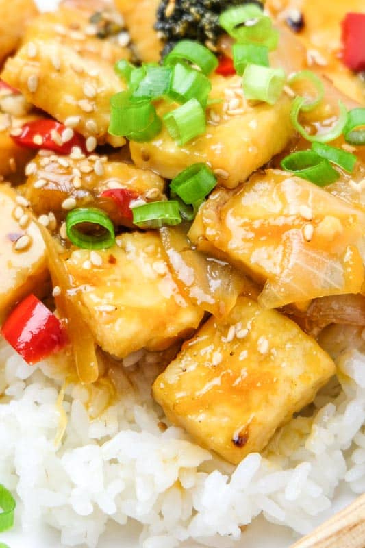 Pieces of tofu with orange sauce and red pepper, green onion, sesame seeds, white rice, onions and orange zest.