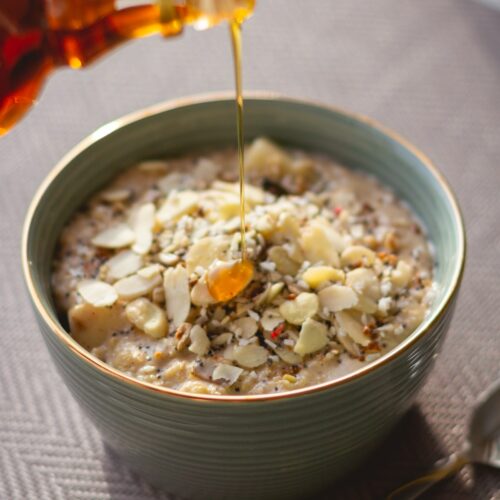 Oatmeal in a bowl with some extra nuts and grains added and maple syrup being poured over.