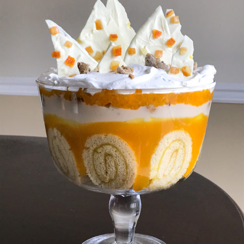 A large glass trifle dish filled with roll cake, orange and white colored layers with pointed shards of almond bark at top.