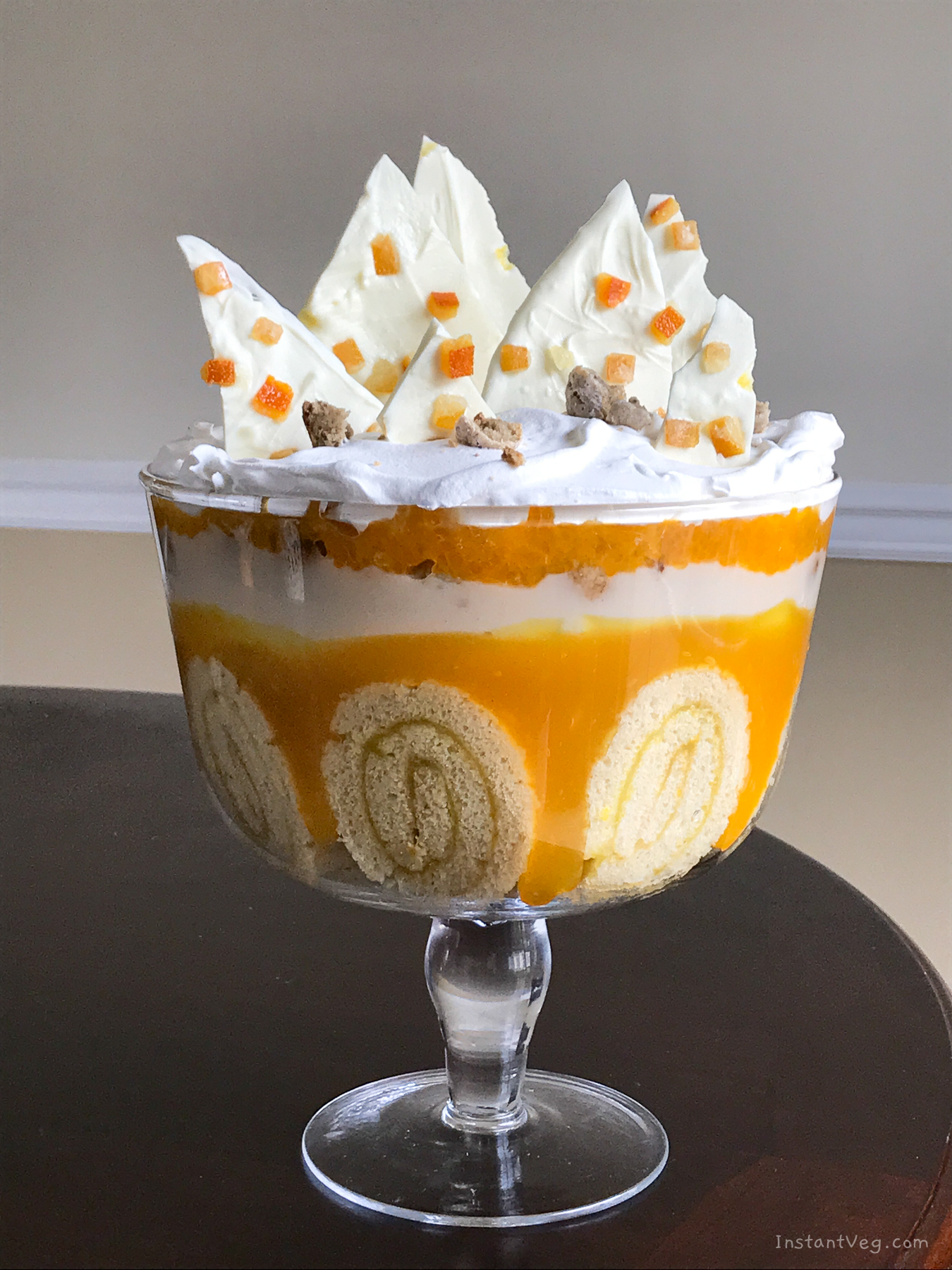 A large glass trifle dish filled with roll cake, orange and white colored layers with pointed shards of almond bark at top.